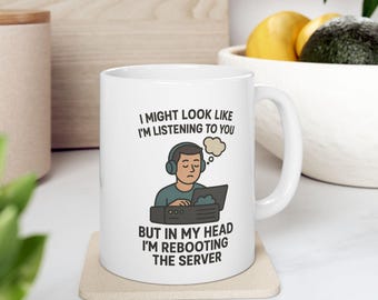 Puede incluir: Taza de cerámica blanca con una ilustración en negro de una persona con auriculares mirando una pantalla de ordenador. El texto en la taza dice "Puede que parezca que te estoy escuchando, pero en mi cabeza estoy reiniciando el servidor."