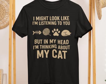 May include: Black t-shirt with the text "I MIGHT LOOK LIKE I'M LISTENING TO YOU BUT IN MY HEAD I'M THINKING ABOUT MY CAT." The shirt has cat-related graphics, including a fishbone, yarn ball, paw print, and a sleeping cat.