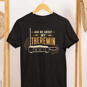 May include: Black t-shirt with a yellow and white graphic. The graphic reads "= ASK ME ABOUT = MY THEREMIN" with an illustration of a theremin instrument. The shirt is hanging on a wooden hanger.