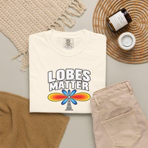 May include: A cream-colored t-shirt featuring the text LOBES MATTER in bold black letters above a graphic of a radio tower with blue, orange, and yellow signal waves. The shirt has a Comfort Colors brand label at the inner neckline.