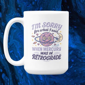 Pode incluir: Caneca de cerâmica branca com uma alça curva. A caneca apresenta o texto "I'M SORRY for what I said WHEN MERCURY WAS IN RETROGRADE" com um design de planeta e lua de desenho animado em roxo, rosa e amarelo.