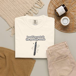 May include: A cream-coloured t-shirt featuring the black text "breathing controls the spaceship sounds" above a graphic of a black electronic wind instrument surrounded by small star icons. The shirt has a crew neckline and a Comfort Colors brand label.