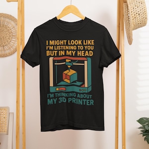 Könnte beinhalten: Schwarzes T-Shirt mit einem Aufdruck einer 3D-Druckers und dem Text "I MIGHT LOOK LIKE I'M LISTENING TO YOU BUT IN MY HEAD I'M THINKING ABOUT MY 3D PRINTER". Das Design verwendet Teal, Orange und Beige.