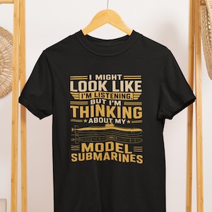 Può includere: Una t-shirt nera a maniche corte con testo dorato che recita I MIGHT LOOK LIKE I'M LISTENING, BUT I'M THINKING ABOUT MY MODEL SUBMARINES, accompagnata da una dettagliata illustrazione grafica dorata di un sottomarino visto di lato e dall'alto.