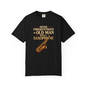 May include: Black t-shirt featuring a gold saxophone graphic and the text "Never Underestimate an Old Man with a Saxophone." The design is in a vintage style. The shirt is made of a soft material.
