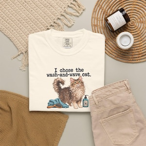 Może przedstawiać: Kremowy t-shirt z grafiką puszystego brązowego kota z kręconą sierścią. Tekst nad kotem brzmi, I chose the wash-and-wave cat. Pod kotem znajduje się niebieski ręcznik, szczotka do włosów oraz mała butelka szamponu dla zwierząt.