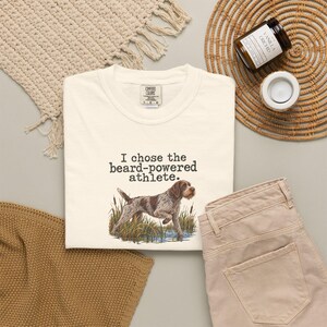 May include: A cream-colored t-shirt featuring a graphic of a brown and white wirehaired pointing dog standing in tall grass and water. Above the dog, the text reads, "I chose the beard-powered athlete." The shirt has a crew neckline and a tag.