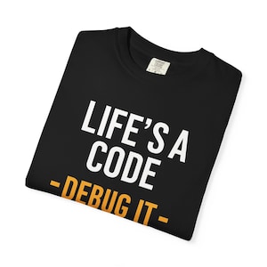 Könnte beinhalten: Schwarzes T-Shirt mit dem Text "LIFE'S A CODE - DEBUG IT" in Weiß und Orange. Das T-Shirt ist gefaltet. Das T-Shirt hat einen Rundhalsausschnitt und kurze Ärmel. Der Text ist ein Wortspiel für Programmierer.