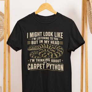 Op de afbeelding: Een zwart t-shirt met korte mouwen en een afbeelding van een opgerolde slang. De tekst luidt: I MIGHT LOOK LIKE I'M LISTENING TO YOU BUT IN MY HEAD I'M THINKING ABOUT MY CARPET PYTHON. Het ontwerp is in lichtbeige kleur op de voorkant gedrukt.