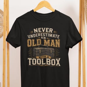 May include: Black t-shirt with the text "NEVER UNDERESTIMATE AN OLD MAN WITH A TOOLBOX" in white and tan. The design includes a toolbox graphic. The shirt is hanging on a wooden hanger.