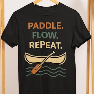 May include: Black t-shirt with the words "PADDLE. FLOW. REPEAT." in orange, teal, and beige. Below the text is a beige canoe with an orange paddle and teal waves. The shirt is hanging on a wooden hanger.