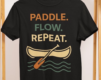 Puede incluir: Camiseta negra con las palabras "PADDLE. FLOW. REPEAT." en naranja, verde azulado y beige. Debajo, una canoa beige con una paleta naranja y olas turquesas. La camiseta está colgada en una percha de madera.
