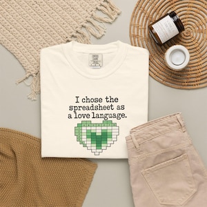 Puede incluir: Una camiseta de color crema con el texto negro "I chose the spreadsheet as a love language." debajo del cual hay un gráfico en forma de corazón compuesto por una cuadrícula de cuadrados de color verde claro y oscuro, que se asemeja a una hoja de cálculo con filas y columnas etiquetadas.