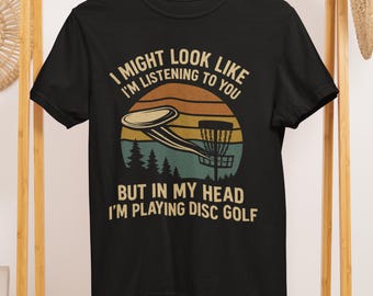 May include: Black t-shirt with a disc golf design. The graphic features a disc, basket, and text that reads "I MIGHT LOOK LIKE I'M LISTENING TO YOU BUT IN MY HEAD I'M PLAYING DISC GOLF". The design is in shades of brown, yellow, and teal.