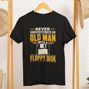 Puede incluir: Una camiseta negra de manga corta con un estampado gráfico en texto blanco y amarillo que dice: NEVER UNDERESTIMATE AN OLD MAN WITH A FLOPPY DISK, acompañado de una ilustración blanca y amarilla de un icono de disquete cuadrado.