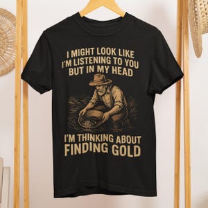Peut inclure: T-shirt noir avec un dessin d'un chercheur d'or et le texte : "I MIGHT LOOK LIKE I'M LISTENING TO YOU BUT IN MY HEAD I'M THINKING ABOUT FINDING GOLD." Le design est en nuances de marron et beige.