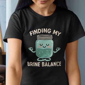 Op de afbeelding: Zwart T-shirt met de tekst "FINDING MY BRINE BALANCE". Het shirt heeft een cartoon illustratie van een pot in een yoga pose, met een lachend gezicht en armen in de vorm van de handen in de mudra gebaar.