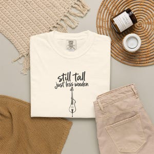 Könnte beinhalten: Ein cremefarbenes T-Shirt mit dem schwarzen Schriftzug "still tall just less wooden" über einer grafischen Illustration eines Saiteninstruments. Das T-Shirt hat ein Comfort Colors Markenetikett am inneren Halsausschnitt und liegt flach.