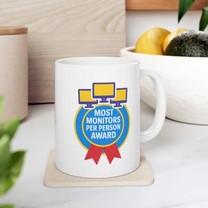 Op de afbeelding: Witte keramische mok met een blauwe cirkel en een rood lint. De cirkel bevat de tekst "Most Monitors Per Person Award" en een afbeelding van vier computermonitoren.