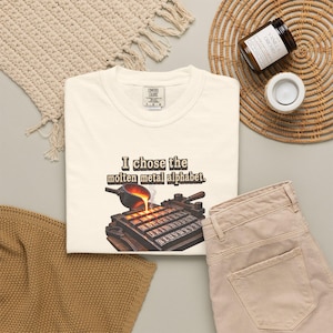 Könnte beinhalten: Ein cremefarbenes T-Shirt mit einer Grafik, die eine Schöpfkelle zeigt, welche geschmolzenes Metall in eine Form in Buchstabenform gießt. Der Text über der Grafik lautet: "I chose the molten metal alphabet." Das T-Shirt hat ein Comfort Colors Etikett.