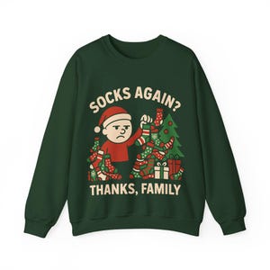 May include: A dark green sweatshirt with a humorous Christmas graphic. The graphic features a cartoon character wearing a Santa hat, surrounded by socks and presents, with the text "SOCKS AGAIN? THANKS, FAMILY."