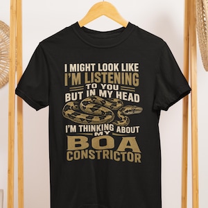 May include: A black short-sleeved t-shirt featuring a graphic of a coiled snake in the centre. The text reads: I MIGHT LOOK LIKE I'M LISTENING TO YOU BUT IN MY HEAD I'M THINKING ABOUT MY BOA CONSTRICTOR. The design is printed in a muted gold colour.