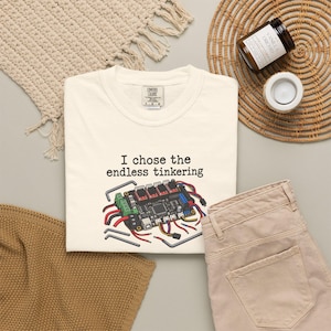 May include: A cream-colored t-shirt featuring a graphic of a 3D printer control board with colorful wires and the text "I chose the endless tinkering" printed in black. The shirt has a Comfort Colors brand label at the inner neckline.