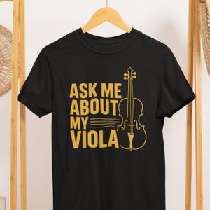 May include: Black t-shirt with the text "ASK ME ABOUT MY VIOLA" in gold lettering. A gold outline of a viola is to the right of the text. The shirt is hanging on a wooden hanger.