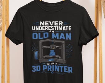 May include: Black t-shirt featuring the text "NEVER UNDERESTIMATE AN OLD MAN WITH A 3D PRINTER" in white and blue. The design includes a 3D printer and a blue filament spool. The shirt is displayed on a wooden hanger.