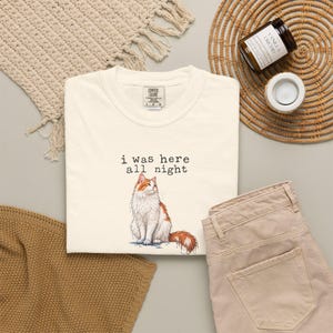 Może przedstawiać: Kremowy t-shirt z grafiką przedstawiającą siedzącego kota o długiej, pomarańczowo-białej sierści. Nad kotem znajduje się napis "i was here all night" czcionką przypominającą maszynę do pisania. Koszulka posiada klasyczny okrągły dekolt pod szyją.