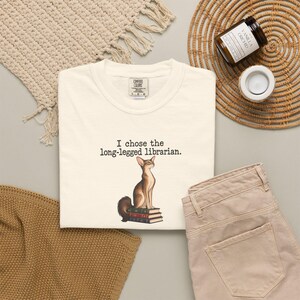 Könnte beinhalten: Ein cremefarbenes Kurzarm-T-Shirt mit einer Grafik einer braunen Katze, die auf einem Stapel von drei Büchern sitzt. Der Text über der Katze lautet: "I chose the long-legged librarian." Das T-Shirt hat einen Rundhalsausschnitt und ein Markenetikett.