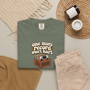 Puede incluir: Una camiseta de color verde oliva con un gráfico de una caja marrón llena de discos de vinilo de colores. El texto sobre la caja dice "one more record won't hurt" en una fuente negrita de color crema con un contorno marrón oscuro.