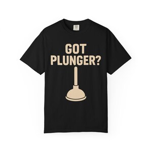 以下が含まれることがあります： 「GOT PLUNGER?」の文字が薄茶色のフォントでプリントされた黒いTシャツ。その下に、薄茶色のラバーカップのイラスト。クルーネックの半袖Tシャツです。