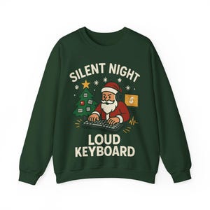 Puede incluir: Sudadera verde oscuro con un gráfico de Papá Noel con auriculares y escribiendo en un teclado. El texto dice "SILENT NIGHT LOUD KEYBOARD" con un árbol de Navidad y copos de nieve.