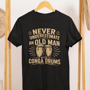 Puede incluir: Camiseta negra con el texto "NUNCA SUBESTIMES A UN ANCIANO CON CONGAS" en color beige. El diseño incluye ilustraciones de congas y líneas decorativas.