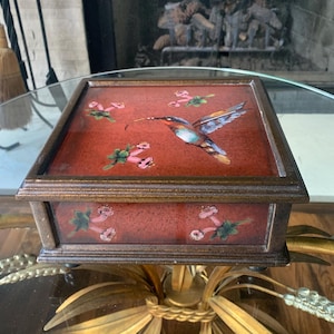 May include: A square, decorative box with a hummingbird design. The box has a red background with a painted hummingbird and floral accents. The box is brown with gold trim and sits on a gold-colored stand.