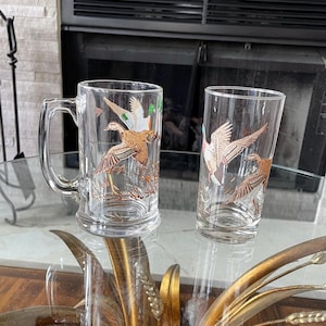 May include: Two clear glass drinking glasses with a gold duck design. One is a handled mug, the other a tall tumbler. The duck design features the birds in flight. The glasses are on a glass table.