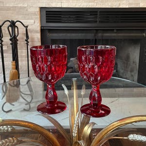 May include: Two ruby red wine glasses with a honeycomb pattern. The glasses have a rounded bowl, a stem, and a circular base. The glasses are displayed on a glass table with a decorative gold base, in front of a fireplace.