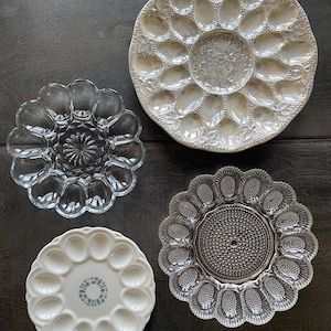 May include: A collection of four egg plates including one iridescent white plate with a floral center, one clear glass plate with a scalloped edge, one white ceramic plate with a central geometric pattern, and one clear glass plate with a textured center.