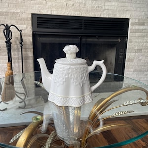 May include: A white ceramic teapot with a floral design and a decorative lid. The teapot is placed on a glass table with a gold-colored metal base. The background includes a fireplace and a brick wall.