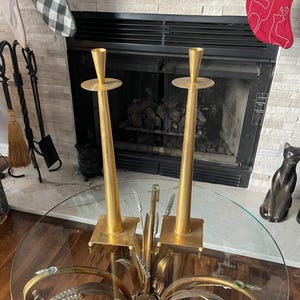 May include: Two gold-colored candle holders with a square base and a tall, slender stem. The candle holders are placed on a glass-topped table with a decorative metal base. A fireplace and Christmas stockings are in the background.