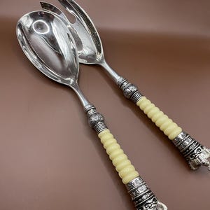 May include: A set of two silver salad servers with ornate handles. The handles are made of a cream-colored material and feature a crown design at the end.