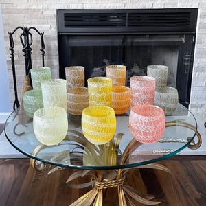May include: A collection of colorful glass votive candle holders on a glass-topped table. The holders are in shades of yellow, orange, pink, green, and brown, with a white web-like design. The table has a gold-colored base.