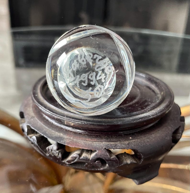 May include: A clear, faceted crystal ball with etched text, resting on a dark brown, carved wooden stand. The ball's interior features intricate details. The stand has a round top and a decorative, carved base.