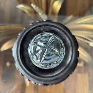 May include: A clear, cut-glass crystal bottle stopper with a starburst design. The stopper is set in a dark, ornate metal base. The background features gold-colored, stylized floral elements.