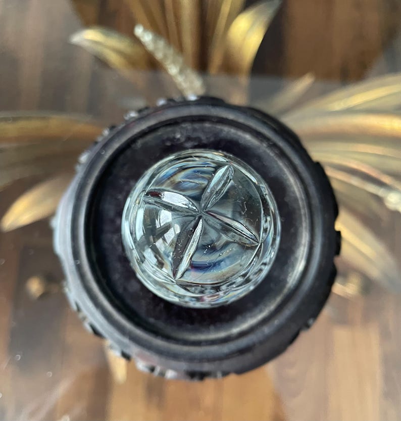 May include: A clear, cut-glass crystal bottle stopper with a starburst design. The stopper is set in a dark, ornate metal base. The background features gold-colored, stylized floral elements.