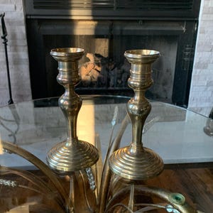 May include: Two brass candle holders with a fluted base and a flared top. The candle holders are displayed on a glass table with a decorative gold base. The background includes a fireplace.