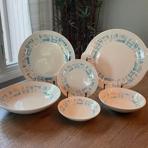 May include: A collection of white ceramic dinnerware with a blue and gray geometric pattern. The set includes plates, bowls, and a serving dish, arranged on a wooden surface. The dishes are displayed on black stands.