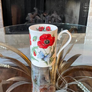 May include: White ceramic mug featuring a vibrant red poppy design with green stems and leaves, and light blue butterflies. The mug has a curved handle and is set on a glass table.