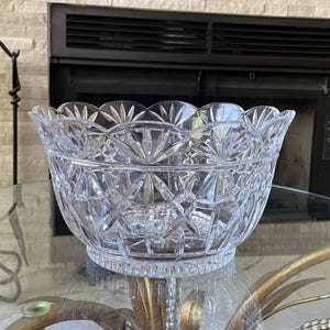 May include: A clear, cut-glass bowl with a scalloped rim and intricate geometric patterns. The bowl sits on a glass table, reflecting light and showcasing its detailed design. The bowl is a decorative piece.
